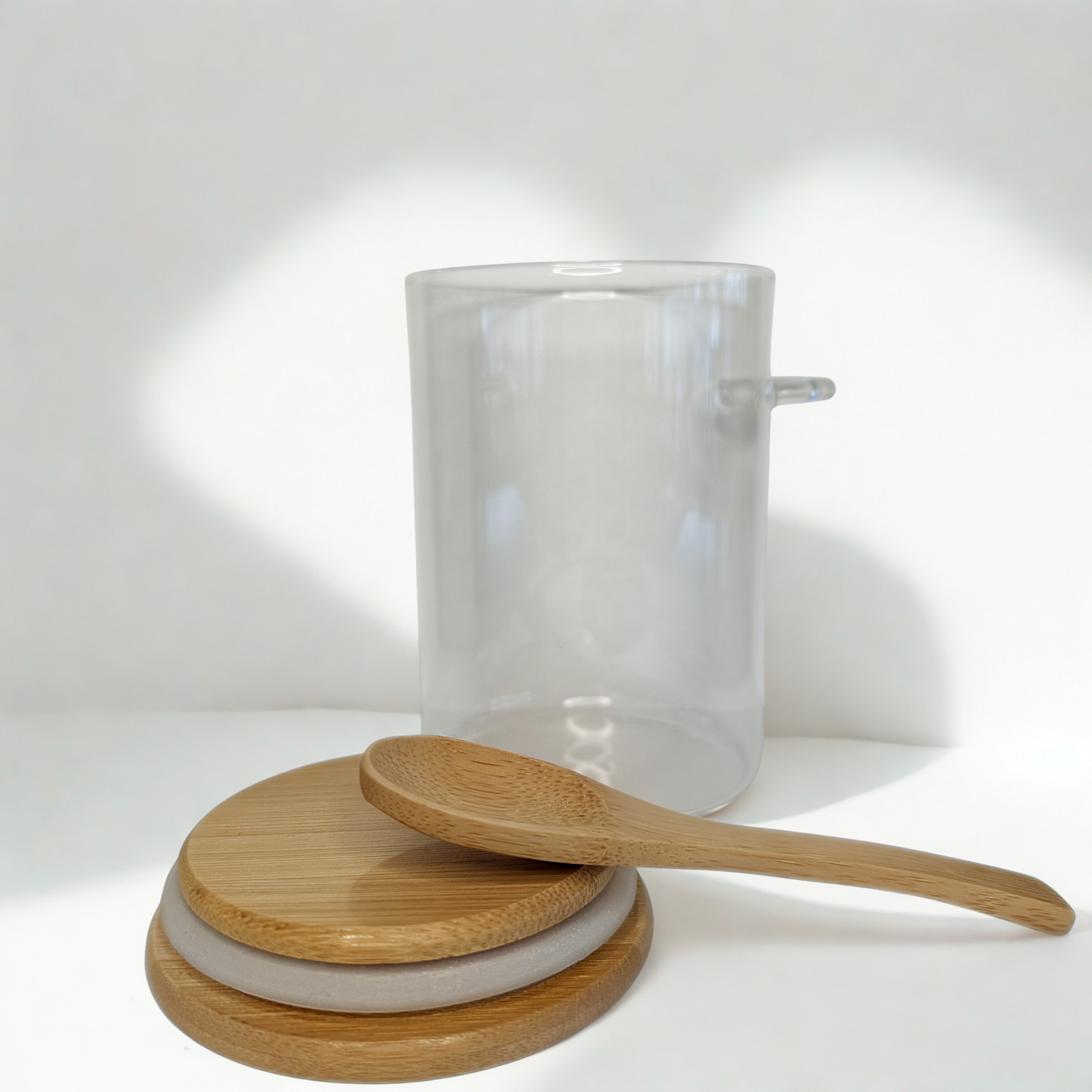 Bamboo lid with bamboo spoon laid on top and an open glass jar behind on a white surface and with background